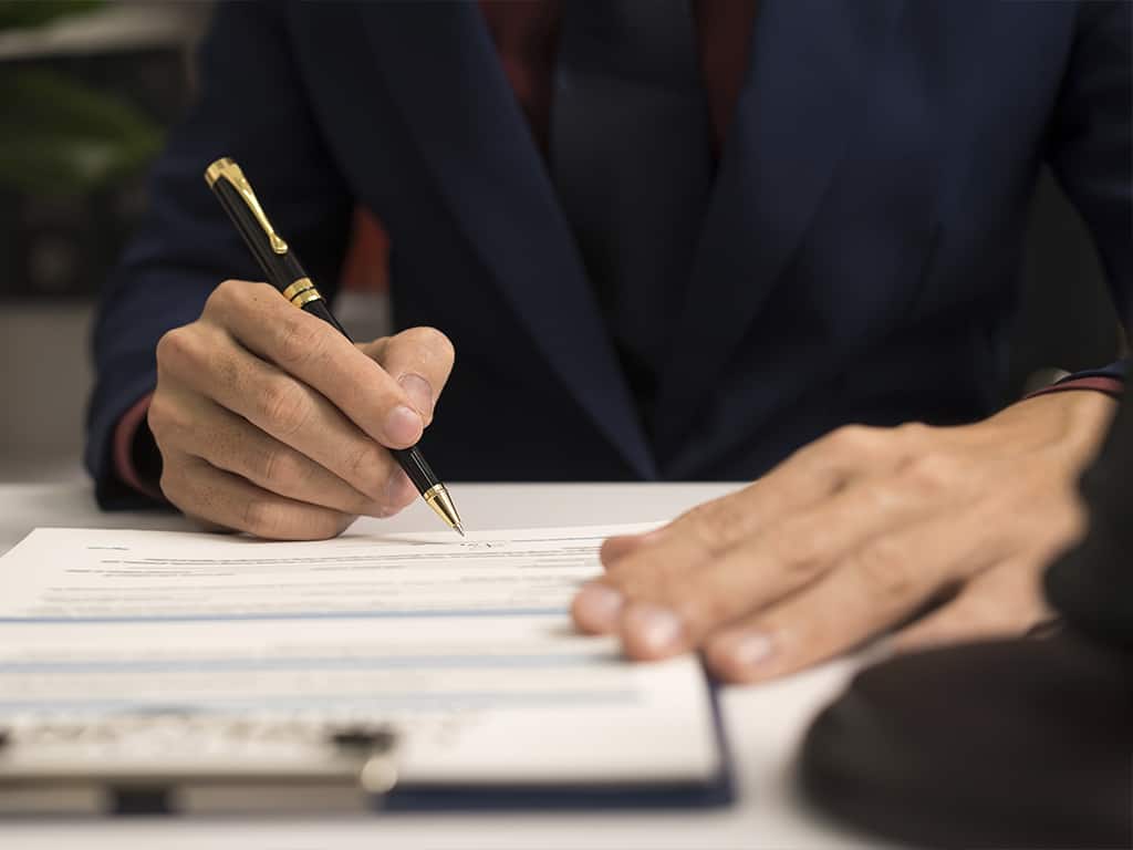 A person in a suit holding a pen, and signing documents on a clipboard